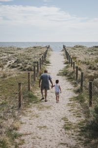 A mature man and young boy walk hand-in-hand along a pathway between two sand dunes towards the ocean.